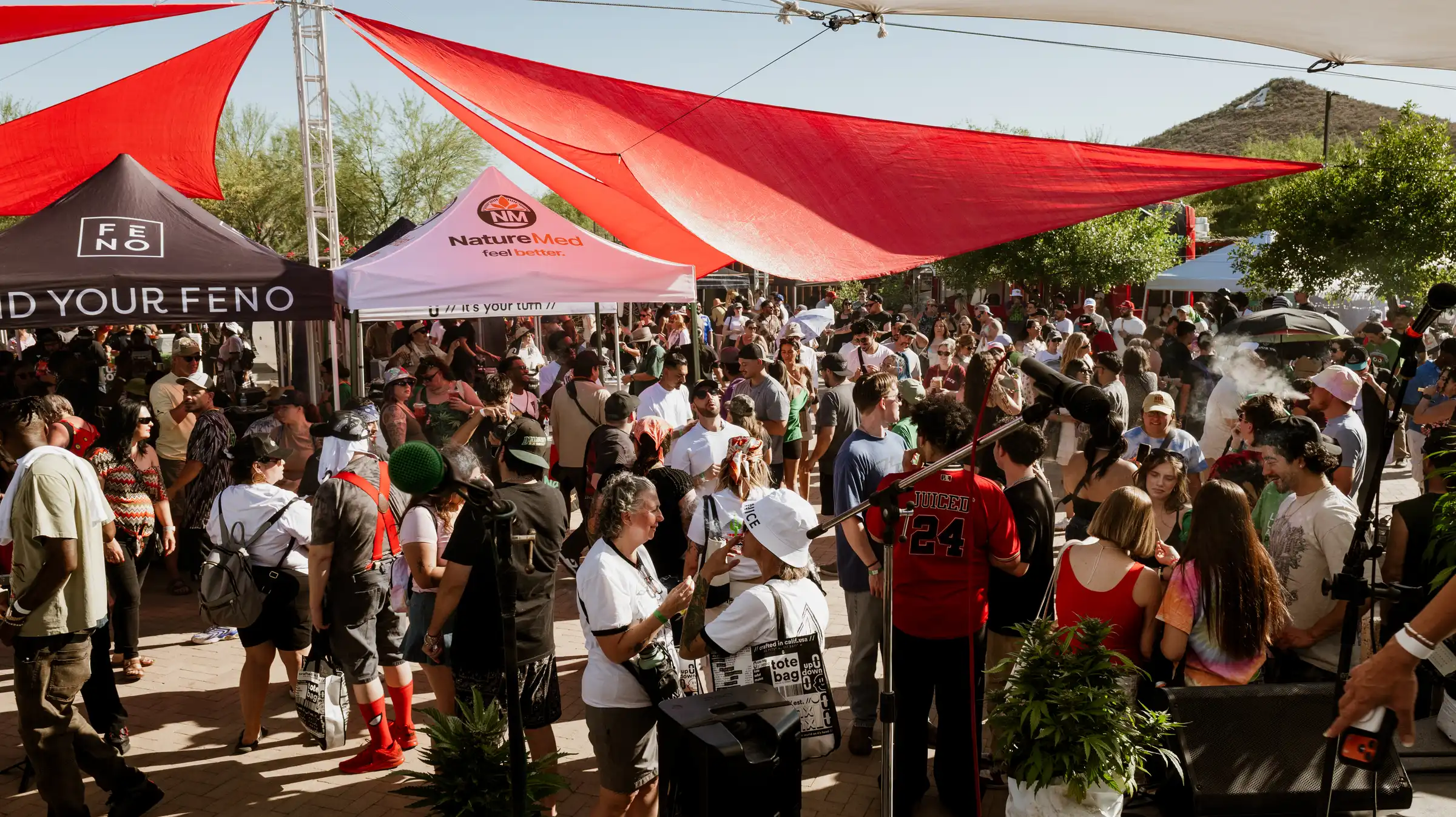 A crowded outdoor cannabis festival with red sail shades and brand tents.